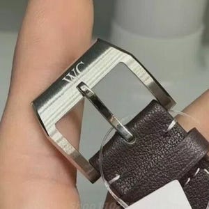 Puede incluir: Primer plano de una hebilla de correa de reloj. La hebilla plateada tiene las letras "WC" grabadas. La correa de cuero marr&oacute;n oscuro est&aacute; cosida con hilo blanco.