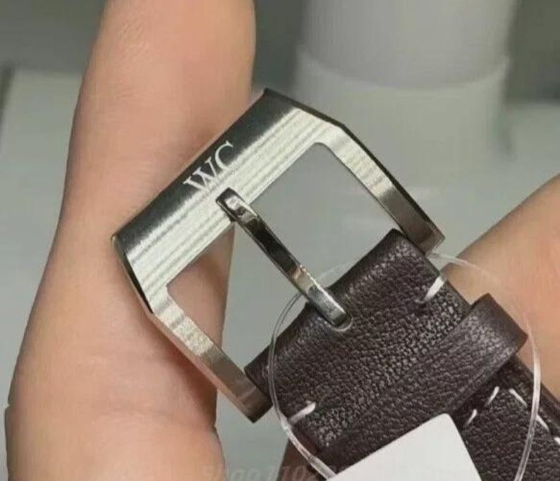 Puede incluir: Primer plano de una hebilla de correa de reloj. La hebilla plateada tiene las letras "WC" grabadas. La correa de cuero marr&oacute;n oscuro est&aacute; cosida con hilo blanco.