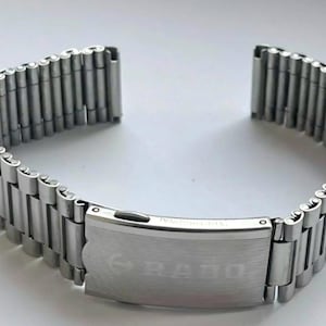 May include: A silver-toned watch band with a folding clasp. The band is made of multiple small, rectangular links. The clasp is engraved with the brand name "RADO" and the words "INTERNATIONAL".