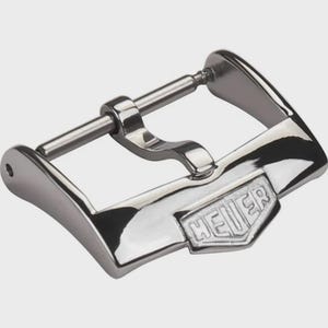 Puede incluir: Hebilla de reloj plateada y brillante, con el nombre de la marca "HEUER" grabado. La hebilla tiene forma rectangular con un diseño curvo y un pasador para asegurar la correa.