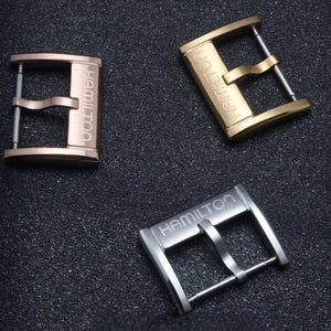 Peut inclure: Trois boucles de montre de couleurs or rose, or et argent. Chaque boucle porte le nom de la marque "HAMILTON" gravé. Les boucles sont présentées sur une surface sombre et texturée.