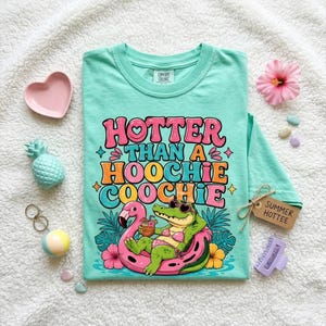 Könnte beinhalten: Ein türkisfarbenes Comfort Colors T-Shirt mit dem Schriftzug "Hotter Than A Hoochie Coochie" in bunten Farben. Die Grafik zeigt ein Alligator mit Sonnenbrille auf einem rosa Flamingo-Schwimmer. Ein Etikett lautet "Summer Hottee".
