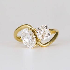 May include: A gold ring featuring two diamonds, one oval and one square-cut. The diamonds are set in a flowing, open design. The ring is set against a white background, highlighting the sparkle of the gemstones.
