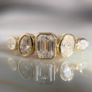May include: A gold ring featuring an emerald-cut diamond center stone, flanked by four smaller diamonds in various shapes. The diamonds are set in a bezel setting, creating a modern and elegant design. The ring is displayed on a reflective surface.