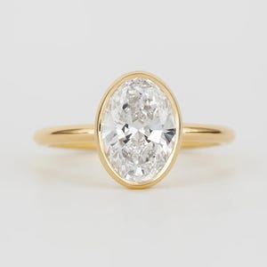 May include: A gold ring featuring a central oval-cut clear gemstone held in a smooth bezel setting. The band is thin, polished, and circular, crafted from yellow gold. The gemstone is faceted, reflecting light with a brilliant, clear appearance.