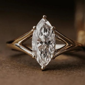 May include: A marquise-cut clear gemstone ring set in a polished gold band. The ring features a split-shank design where the band divides into two distinct arms on each side, meeting at the central prong setting that holds the elongated stone.