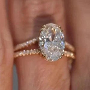 May include: A gold engagement ring with a large oval-cut diamond as the centerpiece. The band is also gold and features smaller accent diamonds. The ring is set on a finger, showcasing its design and sparkle.