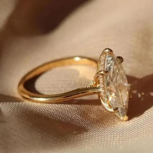 May include: A gold ring with a large, clear, marquise-cut gemstone. The ring's band is a polished gold, and the gemstone is set with small accent stones. The ring is displayed on a neutral-toned fabric.