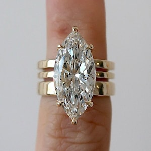 May include: A marquise-cut diamond engagement ring with a gold band, flanked by two additional gold bands. The diamond is set in a gold prong setting, and the bands are smooth and polished. The ring is shown on a finger.