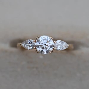 Puede incluir: Un anillo de oro con una gran gema redonda y transparente flanqueada por dos gemas en forma de pera. El anillo está engastado sobre un fondo neutro, resaltando el brillo de las piedras. Una pieza de joyería fina.