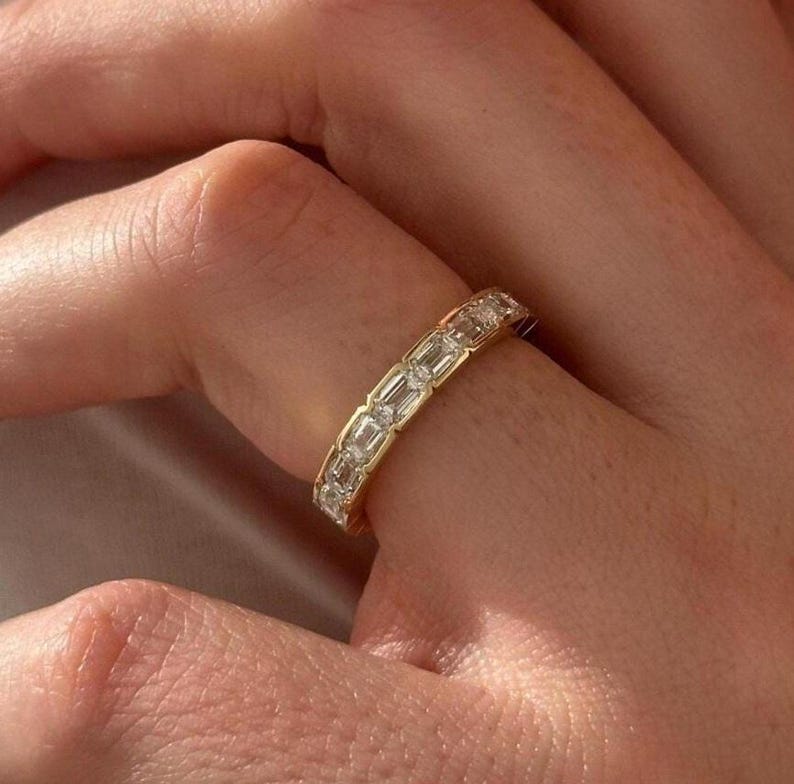 May include: A gold band ring with a series of rectangular-cut diamonds set around the band. The ring is on a finger, and the diamonds sparkle in the light. The ring is a classic and elegant piece of jewelry.
