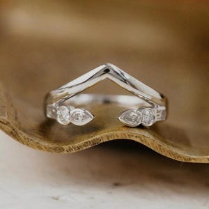 May include: A silver-toned chevron-shaped ring featuring a V-shaped band with an open front design. The ring is accented with small, clear, pear-shaped and round-cut stones embedded along the two ends of the band, creating a symmetrical look.