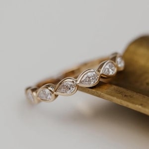 Può includere: Un anello a fascia in oro con una serie di pietre trasparenti sfaccettate a forma di goccia disposte in fila continua. Il metallo ha una finitura lucida e calda, e ogni pietra è fissata in una montatura a castone, creando un design a goccia.