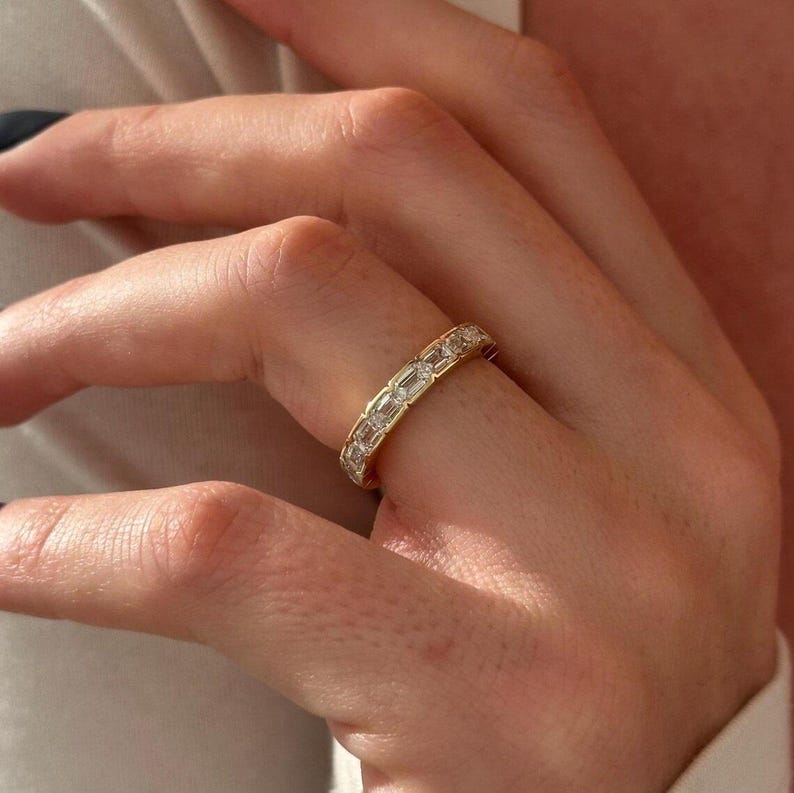May include: A gold band ring with rectangular-cut gemstones set around the entire band. The ring is on a finger, and the gold band contrasts with the skin tone. The ring is a classic design, suitable for various occasions.