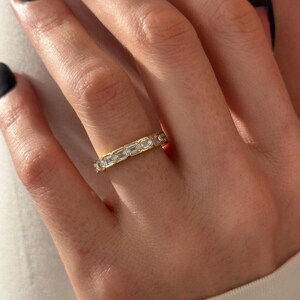 May include: A gold band ring with rectangular-cut gemstones set in a continuous pattern. The ring is shown on a person's finger, with the gemstones reflecting light. The ring is a classic design, suitable for various occasions.