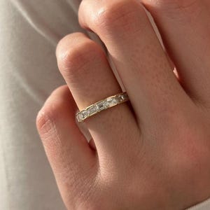 May include: A gold band ring set with rectangular-cut, clear gemstones. The ring is on a finger, with the hand and part of a white garment visible in the background. The ring's design is a continuous pattern of gemstones.