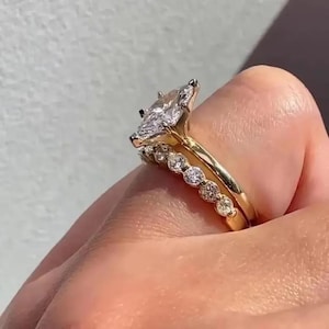 Peut inclure: Une bague de fiançailles en or avec un diamant taille marquise et une bande de petits diamants. La bague est associée à une alliance en or ornée d'une rangée de diamants ronds. Bijoux sur fond neutre.