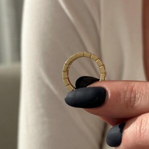May include: A gold ring with a rectangular design is held between fingers with black nail polish. The ring is a continuous band with a repeating pattern of small rectangles. The background is a white shirt.