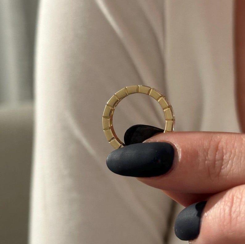 May include: A gold ring with a rectangular design is held between fingers with black nail polish. The ring is a continuous band with a repeating pattern of small rectangles. The background is a white shirt.