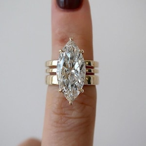 May include: A marquise-cut diamond ring set in yellow gold, featuring a large, elongated center stone held by six prongs. The ring is stacked with two plain, polished yellow gold bands, creating a layered appearance on the finger.