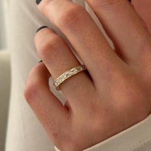 May include: A gold band ring with rectangular-cut gemstones set around the entire band. The ring is on a finger, and the background is a white shirt.