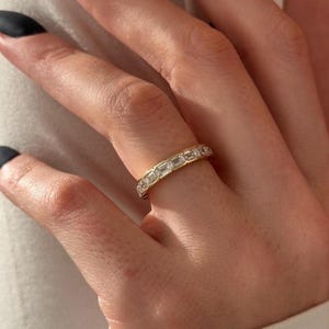 May include: A gold band ring with rectangular-cut clear stones set around the band. The ring is on a finger, with the hand and nails in the frame. The ring is a jewelry item.