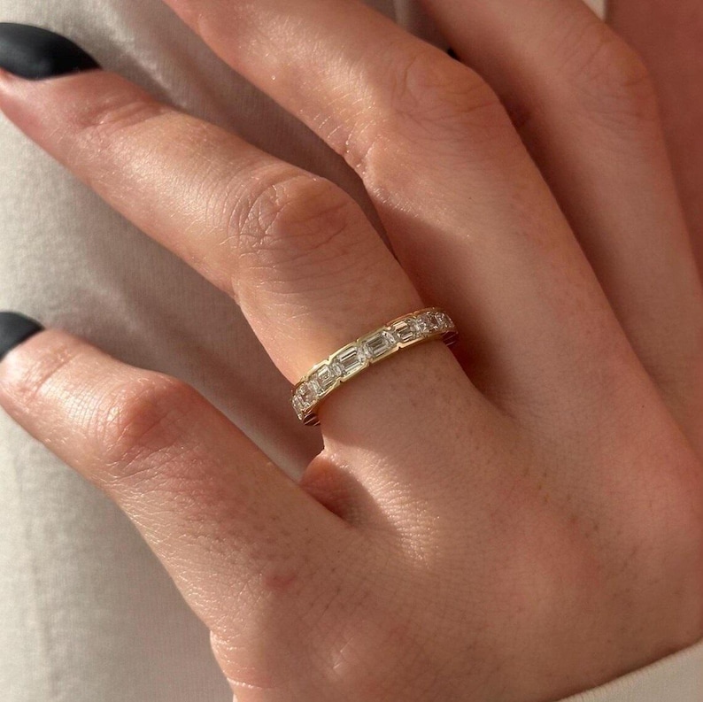May include: A gold band ring with rectangular-cut clear stones set around the band. The ring is on a finger, with the hand and nails in the frame. The ring is a jewelry item.