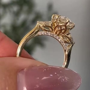 May include: A gold ring featuring a large round center stone held by a floral-inspired setting with petal-shaped side stones. The band is accented with small pavé diamonds along the upper shank, showcasing a polished yellow gold finish.
