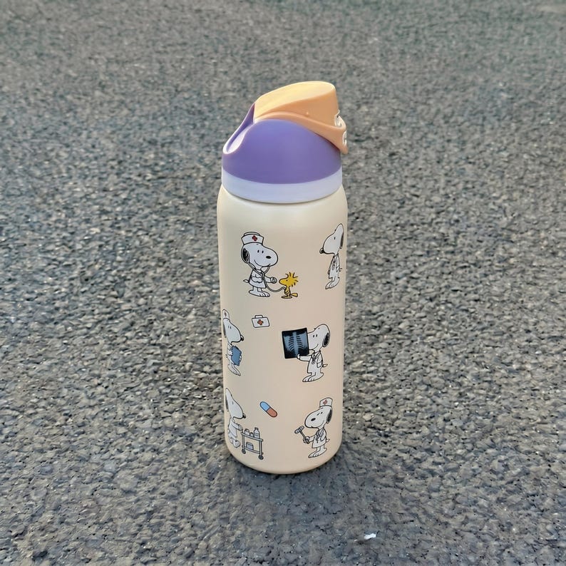 K&ouml;nnte beinhalten: Beige Wasserflasche mit Snoopy-Illustrationen in medizinischen Szenarien. Der Deckel ist lila und orange. Das Design zeigt Snoopy mit Stethoskop, R&ouml;ntgenbildern und anderen medizinischen Bildern. Die Flasche hat eine zylindrische Form.