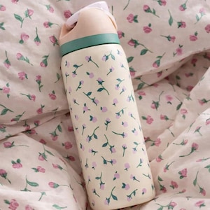 May include: A cream-colored water bottle with a floral pattern of small pink flowers and green stems. The lid is light pink with a green band. The bottle is resting on a floral patterned bedspread.
