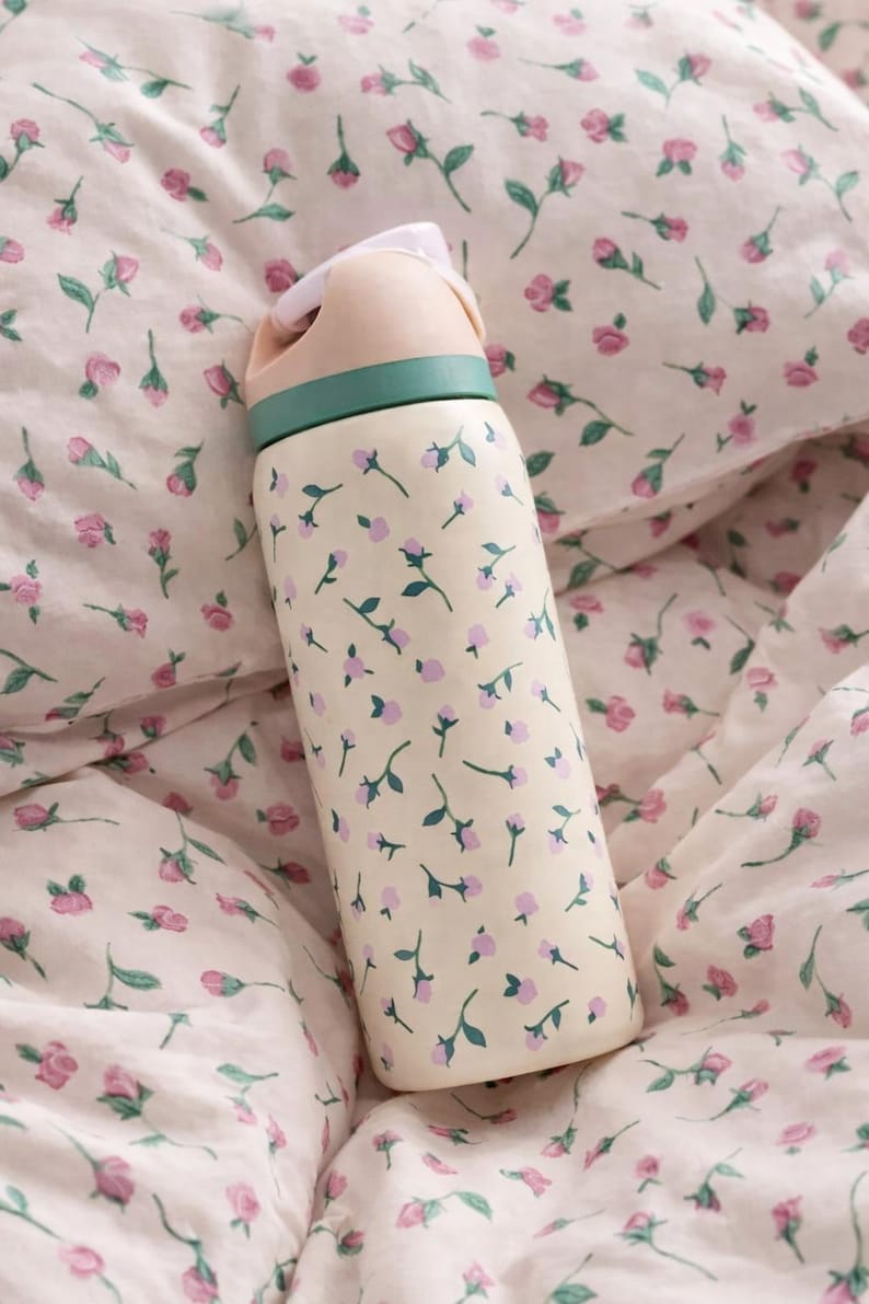 May include: A cream-colored water bottle with a floral pattern of small pink flowers and green stems. The lid is light pink with a green band. The bottle is resting on a floral patterned bedspread.