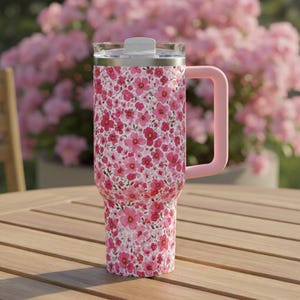 Può includere: Un bicchiere floreale con manico e coperchio rosa. Il bicchiere è ricoperto da un motivo di piccoli fiori rosa su sfondo bianco. Il manico e il coperchio sono rosa tinta unita. Il bicchiere è appoggiato su un tavolo di legno.