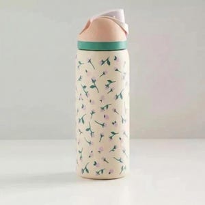 May include: A floral-patterned water bottle with a light beige background and pink and green accents. The bottle features a repeating pattern of small pink flowers and green leaves. The lid is pink and white, with a green band at the base.
