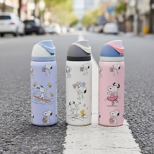 May include: Three stainless steel water bottles with Snoopy designs. The bottles are light blue, white, and pink, each with a different Snoopy graphic. The bottles have a flip-top lid and are standing on a gray road.