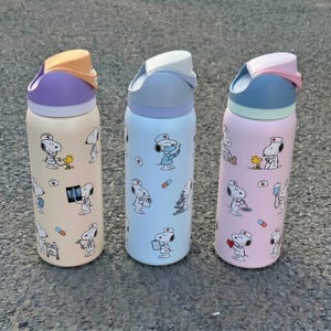 K&ouml;nnte beinhalten: Drei isolierte Wasserflaschen mit Snoopy-Designs. Jede Flasche hat ein anderes Farbschema: Beige mit lila und orangefarbenen Akzenten, hellblau mit wei&szlig;en Akzenten und rosa mit blauen Akzenten. Die Flaschen sind mit Snoopy in verschiedenen medizinischen Illustrationen verziert.