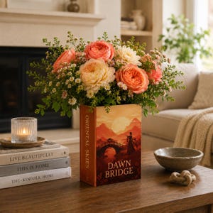 Può includere: Una composizione floreale con rose color pesca e crema e verde in un vaso a forma di libro. Il vaso è intitolato "Dawn Bridge" e "Oriental Skies". La composizione è su un tavolo di legno con altri oggetti decorativi.