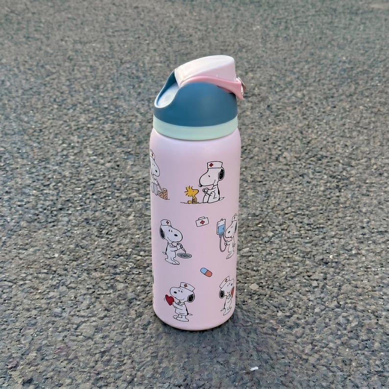 K&ouml;nnte beinhalten: Eine rosa isolierte Wasserflasche mit Snoopy in Krankenschwester-Uniform und medizinischen Illustrationen. Die Flasche hat einen blau-wei&szlig;en Deckel mit rosa Verschluss. Zum K&uuml;hlen von Getr&auml;nken.