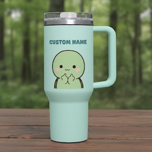 May include: A light teal insulated tumbler with a handle, featuring the text "CUSTOM NAME" above a cartoon character giving the middle finger. The tumbler has a clear lid and a silver rim.