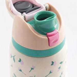 May include: A floral-patterned water bottle with a flip-top lid. The lid is a combination of pink, green, and beige. The bottle's body is cream-colored with pink floral accents. The bottle has a pink latch to secure the lid.