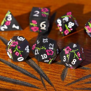 May include: A set of black polyhedral dice with white numbers and pink floral designs. The dice are arranged on a wooden surface, showcasing various shapes, including a D4, D6, D8, D10, D12, and D20.