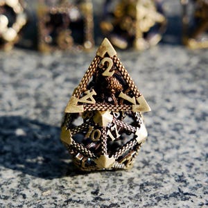 May include: A bronze-colored, geometric D20 die with intricate openwork and raised numbers. The die has a pyramid-shaped top with the number 2 visible. The die is designed for tabletop role-playing games.