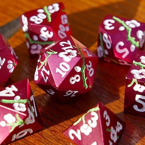 May include: A collection of burgundy polyhedral dice with white floral accents and white numbering. The dice are arranged on a wooden surface. The dice are for role-playing games.