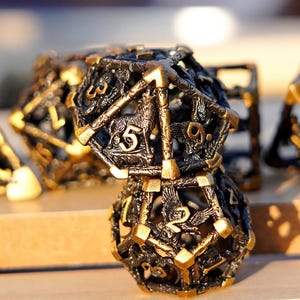 May include: Close-up of two ornate, black and gold D20 dice. The dice feature intricate designs with animal and number details. The dice are made of metal and have a vintage, fantasy aesthetic. Other dice are visible in the background.