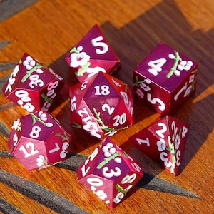 May include: A set of seven burgundy dice with white numbers and floral accents. The dice are arranged on a wooden surface, showcasing their various shapes and sizes, including a D20, D6, and D4.