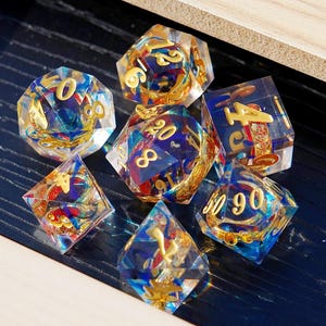 Puede incluir: Un conjunto de siete dados poliédricos con cuerpos de resina transparente, números dorados y elementos decorativos. Los dados tienen acentos azules, rojos y dorados, creando una apariencia vibrante y detallada. Ideal para juegos de mesa.