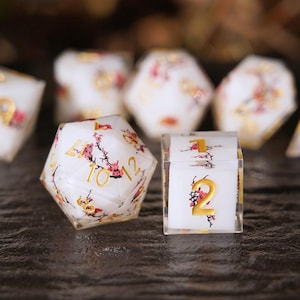 May include: A collection of white polyhedral dice with gold numbering and delicate pink floral designs. The dice are arranged on a dark surface, showcasing their intricate details and artistic flair, perfect for tabletop gaming.