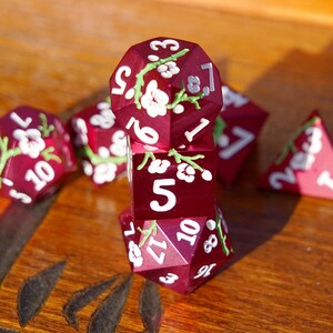 May include: A collection of burgundy polyhedral dice with white floral designs and white numbering. The dice are arranged on a wooden surface, with some dice stacked and others scattered. The dice are for tabletop games.