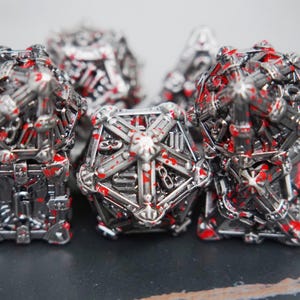 May include: A set of multi-faceted metal gaming dice featuring a complex, cage-like structural design in a metallic silver finish. Each die is accented with irregular, bright red paint splatters across the surface, highlighting the intricate geometric edges.