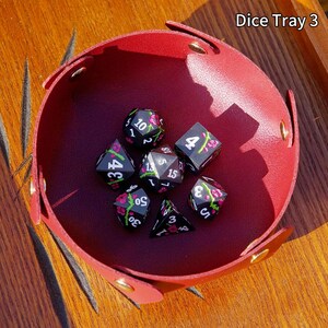 May include: A red leather dice tray with gold rivets holds seven black dice. Each die features white numbers and pink floral designs. The text "Dice Tray 3" is visible in the upper right corner.