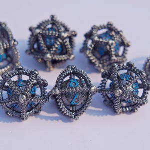 Puede incluir: Un conjunto de dados plateados con un diseño mecánico detallado y detalles en azul. Los dados presentan números azules visibles y vienen en varias formas. Ideales para juegos de mesa, estos dados tienen una apariencia compleja e intrincada.